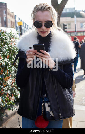 Londres - Février 2017 : taille de la femme portant des lunettes de soleil et un collier blanc veste en fourrure à l'aide de l'extérieur de l'Écosse Pringle smartphone show, London Fashion Week, jour 4. Banque D'Images