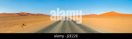 L'interminable route menant hors de Sossusvlei, dans le désert de Namibie - dunes de sable orange vif et le tarmac noir contre le ciel bleu. Banque D'Images