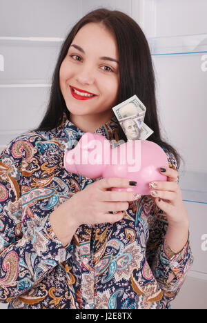 Young Beautiful woman standing with piggy bank (de l'argent fort) sur l'arrière-plan réfrigérateur Banque D'Images