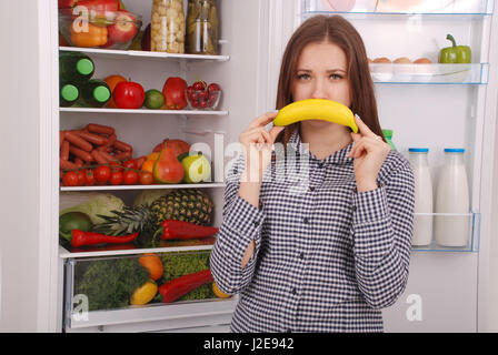 Une jeune fille peut contenir jusqu'à la banane de sa bouche, imitant un sourire. Belle jeune fille près du réfrigérateur. Banque D'Images