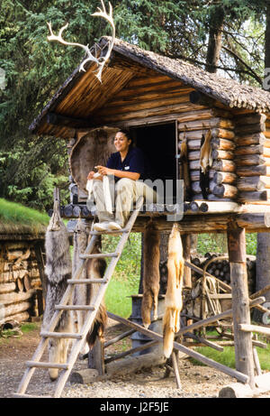 L'Athabaskan femme est assise sur la plate-forme d'un journal alimentaire traditionnellement construit un abri de stockage, appelé cache, situé à l'intérieur d'un camp de chasse et pêche. Village de Chena, Alaska Banque D'Images