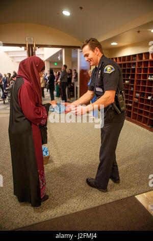 Le port d'un hijab ou foulard et des vêtements traditionnels, une femme musulmane donne aux revêtements de chaussures de protection à un chef de la police locale avant qu'il se joint à la Congrégation pour les services religieux à Anaheim, CA, une mosquée. Des armoires de rangement chaussures Note en arrière-plan. Banque D'Images