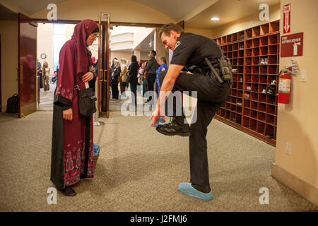 Le port d'un hijab ou foulard et des vêtements traditionnels, une femme musulmane donne aux revêtements de chaussures de protection à un chef de la police locale avant qu'il se joint à la Congrégation pour les services religieux à Anaheim, CA, une mosquée. Des armoires de rangement chaussures Note en arrière-plan. Banque D'Images