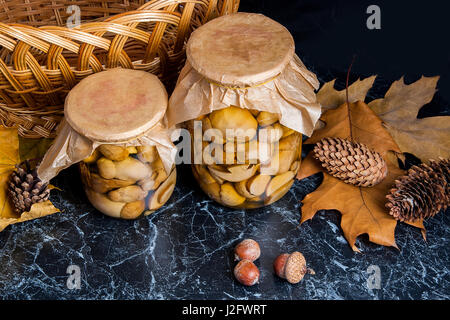 Deux pots en verre avec des champignons marinés sur noir en arrière-plan. Panier en bois jaune sur fond de retour, plusieurs feuilles de chêne, de glands et de sapin Banque D'Images
