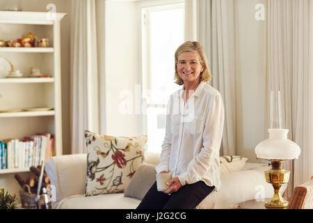 Senior woman looking détendu et heureux dans son salon à la maison, assis sur le bord du bras de son canapé tenant une tasse de café Banque D'Images
