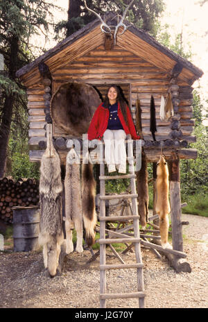 L'Athabaskan Teenage girl se trouve sur la plate-forme d'un journal alimentaire traditionnellement construit un abri de stockage, appelé cache, avec des fourrures animales et une peau de castor. Situé à l'intérieur d'un camp de chasse et pêche. Village de Chena, Alaska Banque D'Images