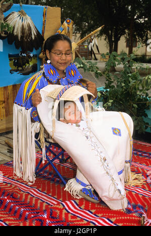 Native American baby avec lacets à l'intérieur d'un porte-bébé traditionnel willow est pris en charge en position verticale par sa sœur qui porte une robe perlée, cape et cheveux. Banque D'Images