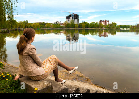 Belle jeune femme assise sur la banque du grand lac à l'horizon de la ville moderne de bâtiments en construction Banque D'Images