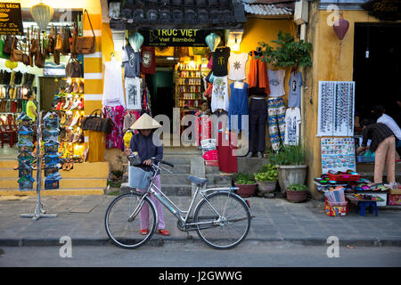 HOI AN, VIETNAM - 15 mars 2014 : femme avec son vélo en Chapeau conique traditionnel sur le marché de la rue de l'ancienne ville de Hoi An, Patrimoine Mondial de l'UNESCO Banque D'Images