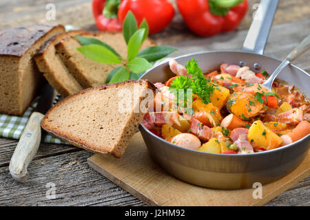 Goulasch de pommes de terre chaudes avec du bacon et saucisses de Vienne servi dans une poêle de fer avec de délicieux pain de campagne au four sombre sur une vieille table en bois Banque D'Images