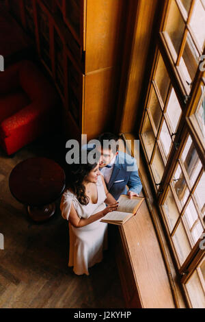 Jeune couple embracing in bibliothèque près de la fenêtre. Charmante épouse penche à son beau palefrenier, holding book Banque D'Images