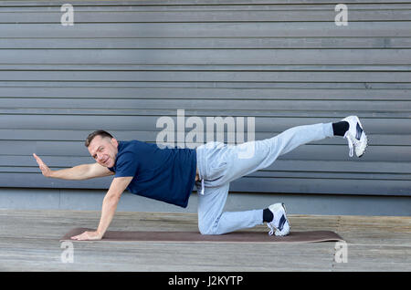 Homme adulte stretching en planche avec une jambe et une main posée et à la recherche à l'appareil photo, vue de côté à l'arrière-plan gris with copy space Banque D'Images