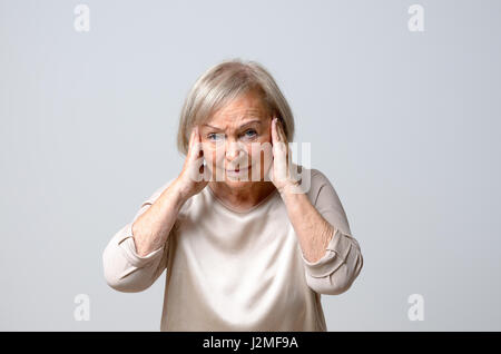 Hauts gray-haired woman touching her temples avec ses deux mains à sa tête, malheureusement looking at camera comme si souffrant de maux de tête, se tenant contre Banque D'Images