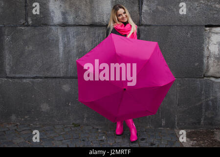 Très belle jeune femme blonde et heureuse avec parapluie colorés sur la rue. Fille dans une écharpe rose vif, des bottes en caoutchouc et parapluie sur le mur de pierre grise de l'arrière-plan Banque D'Images