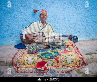 Un musicien de rue joue un sintir le long d'une route à Chefchaouen, Maroc, avec ses murs bleu derrière. Banque D'Images