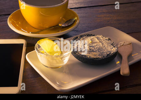 Charbon demi-bagel avec fromage à la crème sur spreaded plaque blanche, fromage à la crème dans la coupe claire avec une tranche de citron, une tasse à café vide avec cuillère en argent, ce Banque D'Images