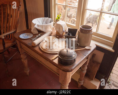 Une gamme d'ustensiles de cuisine anciens et des pots et des assiettes et des couteaux et des fourchettes sur une belle table en bois près d'une fenêtre sur une journée ensoleillée Banque D'Images