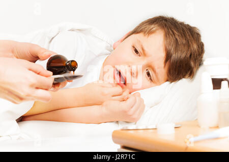 Enfant malade boy laying in bed et de pleurer alors que mère verser le sirop de toux de bouteille avec copie à l'espace-cuillère de dosage Banque D'Images