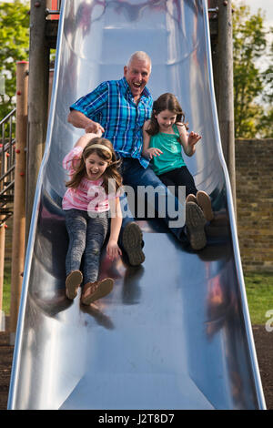 Portrait vertical d'un grand-père glissant sur une diapositive avec ses petits-enfants à une aire de jeux. Banque D'Images