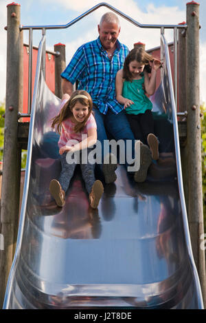 Portrait vertical d'un grand-père glissant sur une diapositive avec ses petits-enfants à une aire de jeux. Banque D'Images