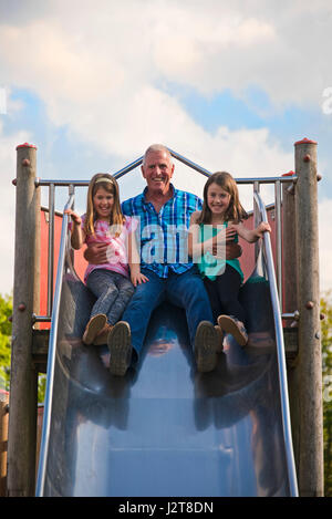 Portrait vertical d'un grand-père glissant sur une diapositive avec ses petits-enfants à une aire de jeux. Banque D'Images