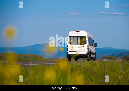 Transport de fret et de passagers van blanc sur la route des fleurs et l'herbe jaune floue à l'avant et d'arbres, montagnes et ciel derrière Banque D'Images