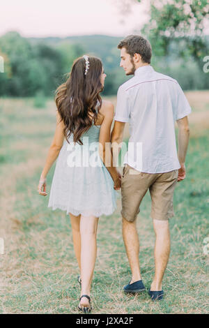 Heureux couple holding hands marcher dans un pré et regarder les uns les autres avec amour. Vue arrière. Belle journée d'été Banque D'Images