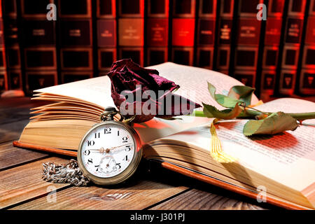 Une rose rouge fanée par les pages d'un vieux livre jauni par le temps et une ancienne montre de poche sur une vieille table en bois. Banque D'Images