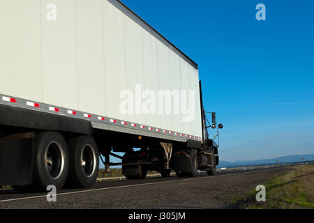 Gros camion semi sombre et blanc sur la route avec remorque,pont,arbres et montagnes et sur fond de ciel bleu Banque D'Images