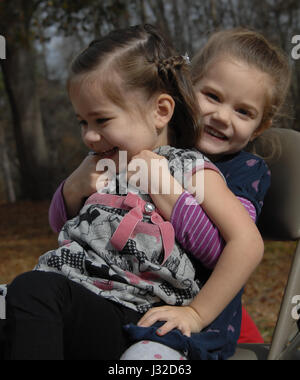 Deux petites filles rire et glousser comme on s'assoit sur les genoux de l'autre. Ils sont assis tous les deux à l'extérieur dans une chaise pliante en métal. Banque D'Images