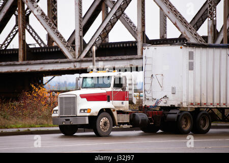 Vieux gréement puissant grand bonnet blanc semi-camion avec une remorque en vrac, couverts de tharp noir sur city road en vertu de l'ancien pont de chemin de fer Banque D'Images