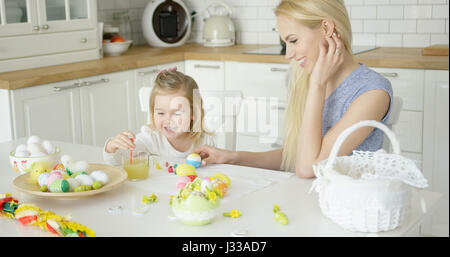 Smiling young woman helping petite fille à la couleur des oeufs de Pâques tout en restant assis à la table de cuisine. Banque D'Images