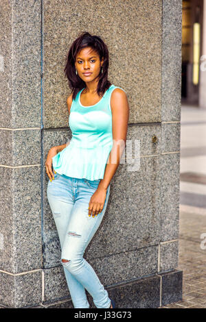 Girl Relaxing extérieur. Débardeur vert vêtu à la mode, jeans, pendants d'oreille, young African American Woman standing by wall à New York, se détendre. Banque D'Images