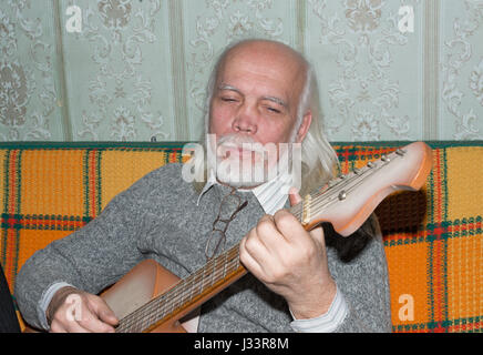 Portrait demi-longueur de pygargues à tête homme âgé avec de longs cheveux gris et sa barbe en gris chandail tricoté s jouer de la guitare assis sur le canapé. Banque D'Images