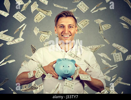 Happy man with piggy bank célèbre son succès sous la pluie tomber de l'argent dollar bills billets isolé sur fond de mur gris. La réussite financière Banque D'Images