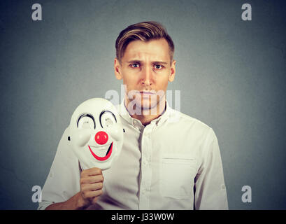 Jeune homme triste avec masque clown heureux isolé sur fond de mur gris. Les émotions humaines Banque D'Images