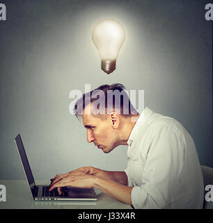 Portrait young man working on computer sitting at desk avec ampoule au-dessus de sa tête isolé sur fond de mur gris. Banque D'Images
