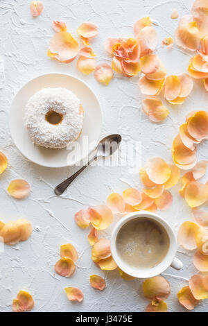 Tasse de café et un beigne sur fond texturé blanc avec des pétales de rose. Petit-déjeuner romantique Banque D'Images