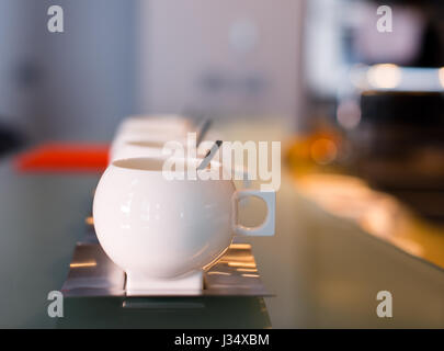 Porcelaine blanche tasse en acier inoxydable contemporaine avec soucoupes et cuillères sur comptoir de verre et fond flou avec taches claires Banque D'Images