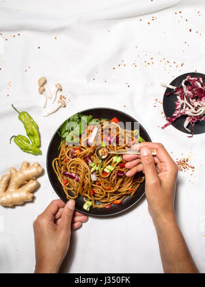 Passage tiré d'un woman's hands à la mise au point d'un sauté de nouilles, avec d'autres ingrédients sur une nappe blanche Banque D'Images