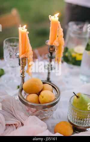 Pique-nique, nourriture, maison de vacances concept - table de pique-nique couverte de fruits, de brûler des bougies dans les chandeliers, un plat d'abricots, poires, un pichet de limonade, des gouttes de cire des bougies, selective focus. Banque D'Images