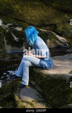 BOLTON ABBEY, UK - 8 avril 2017. Fille aux cheveux bleus s'asseoir sur des pierres à la SRCFA au Yorkshire. Regarder la rivière Wharfe passant entre rock Banque D'Images