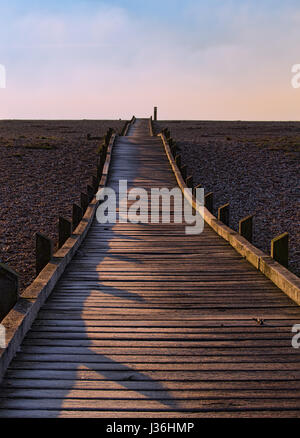 Une petite promenade en bois qui s'étend de la caméra à l'horizon sur une plage de galets en basse lumière directionnelle avec ciel bleu et de nuages au-delà. Banque D'Images