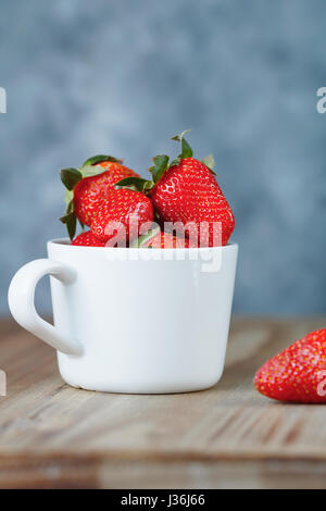 Mug blanc avec des fraises fraîches sur une table en bois contre un mur bleu. Banque D'Images