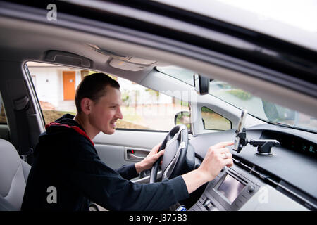 Un jeune conducteur avec un sourire et bonne humeur règle la navigation sur écran tactile sur téléphone mobile tout en restant assis derrière le volant d'une voiture de luxe moderne t Banque D'Images