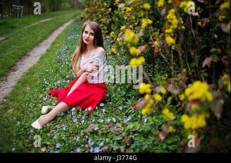 Portrait de sitiing belle fille avec lèvres rouge à fleur de printemps sur l'herbe du jardin de fleurs, l'usure de robe rouge et chemise blanche. Banque D'Images