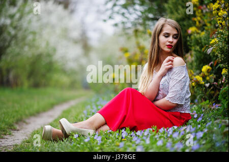 Portrait de sitiing belle fille avec lèvres rouge à fleur de printemps sur l'herbe du jardin de fleurs, l'usure de robe rouge et chemise blanche. Banque D'Images