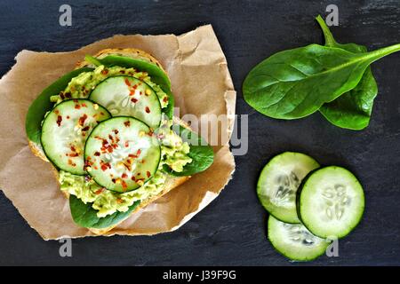 Toast à l'avocat avec le concombre, les épinards et le pain de grains entiers sur papier contre un arrière-plan en ardoise foncé Banque D'Images