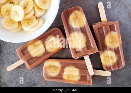 Sucettes glacées banane chocolat dans un cluster de tranches de banane sur fond gris rustique Banque D'Images