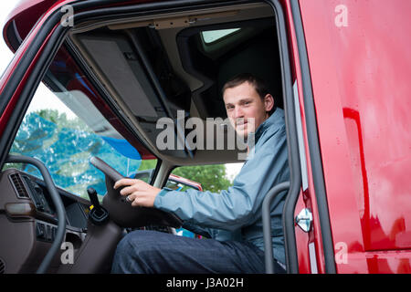 Chauffeur de camion heureux assis dans la cabine de camion rouge derrière le volant et à la recherche à travers la porte ouverte. Le camion semi moderne est situé sur le parking gratuit Banque D'Images